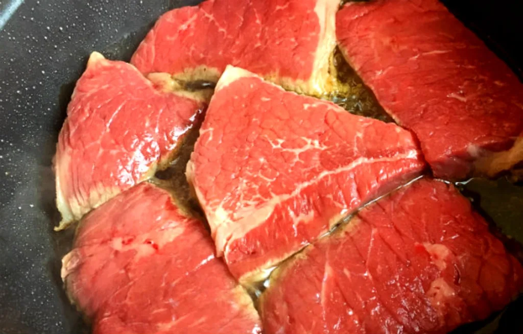 Round steak pieces searing in a skillet to a deep golden brown crust before baking, an important step for rich flavor