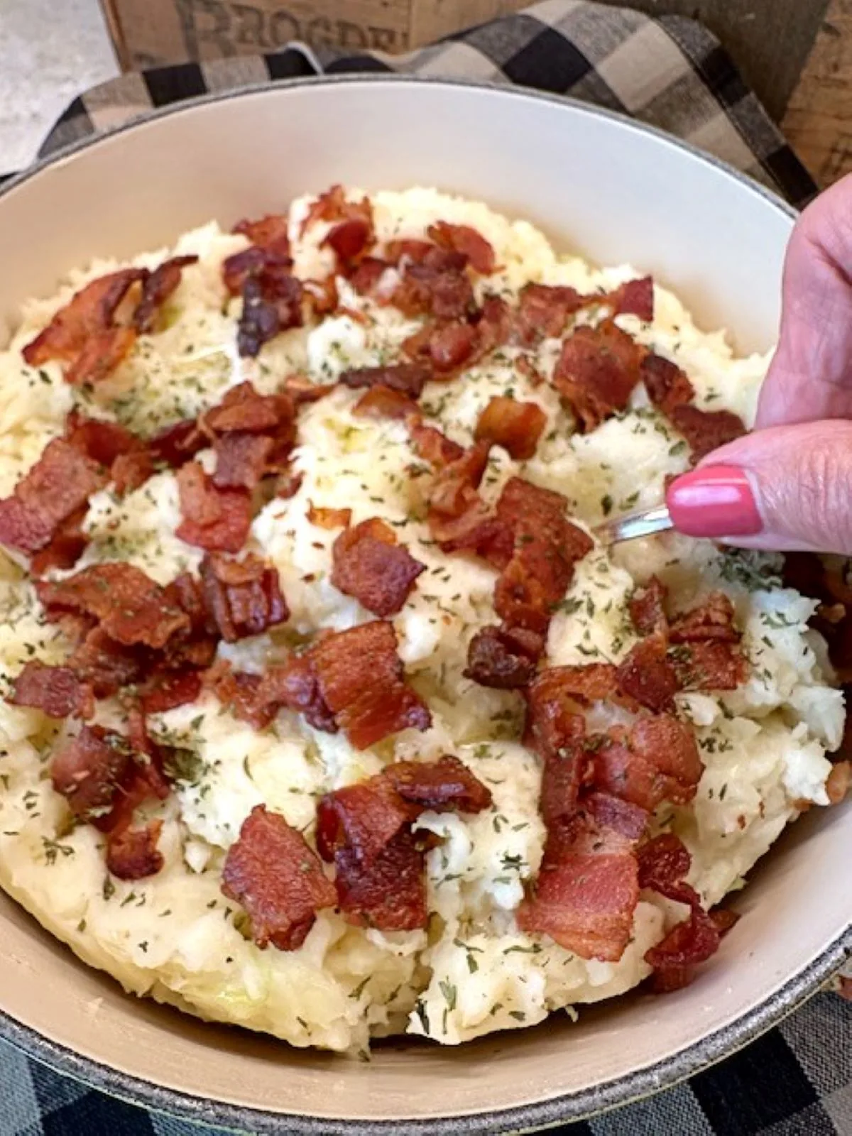 Hand serving creamy Irish Colcannon topped with crisp bacon and herbs in a farmhouse dish on a plaid cloth.